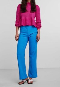 Pull peplum rose en tricot avec des manches longues associé à un pantalon ample bleu vif. Les chaussures présentent des sandales à lanières blanches et noires.