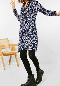 Kobieta ubrana w niebieską, czarną i białą sukienkę w wzory, czarne rajstopy i czarne buty na sznurówki, stojąca obok jasnego drewnianego krzesła.