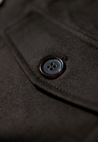 Nahaufnahme eines schwarzen Knopfes, der auf dunklem Stoff mit sichtbaren Nähten und strukturierter Webart angenäht ist.