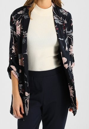 Frau trägt einen cremefarbenen, gerippten Rollkragenpullover, kombiniert mit einer schwarzen, mit Blumenmuster versehenen, offenen Strickjacke und schwarzen Hosen.