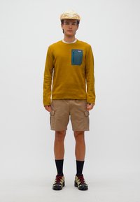 Νεαρός άνδρας που φοράει πουλόβερ μουσταρδί με τσέπη στο στήθος σε βεραμάν χρώμα, μπεζ cargo σορτς, μαύρες κάλτσες, ορειβατικά παπούτσια και μπεζ καπέλο με κάλυμμα για τα αυτιά.