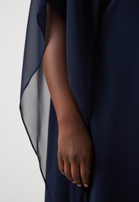 Tissu fluide bleu marine avec des accents transparents. Une main de teinte de peau foncée est partiellement visible, tenant le vêtement, mettant en valeur la texture.