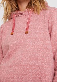 Close-up of person wearing textured pink hoodie with velvet drawstrings and small brown branded tags.