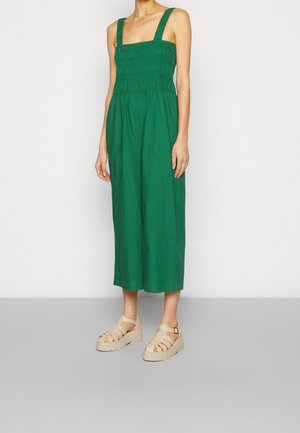 Vrouw draagt een mouwloze groene midi-jurk met brede bandjes en beige plateau sandalen, staand tegen een effen lichte achtergrond.