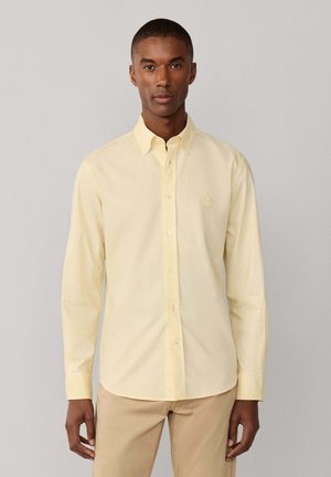 Hombre con una camisa de manga larga abotonada de color amarillo pálido y pantalones beige, de pie contra un fondo liso de color gris claro.