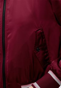 Gros plan d'une veste bordeaux montrant une fermeture éclair noire, une poche boutonnée en biais et un poignet rayé sur la manche.