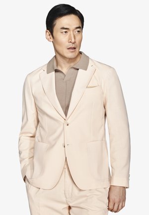 Hombre con traje beige claro y camisa taupe con cuello, con las manos en los bolsillos, mirando hacia su derecha sobre fondo blanco.