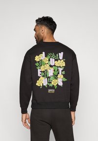 Sudadera negra con un diseño floral que presenta flores amarillas y un texto en tonos pastel que dice "JUVENTUS" en la parte trasera. Puños y dobladillo acanalados.