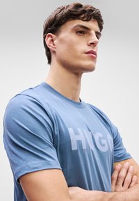 Μπλε βαμβακερό T-shirt με μεγάλη, ανοιχτό μπλε εκτύπωση "HUGO" στο μπροστινό μέρος. Κοντά μανίκια και στρογγυλή λαιμόκοψη. Λείος υφασμάτινος. 