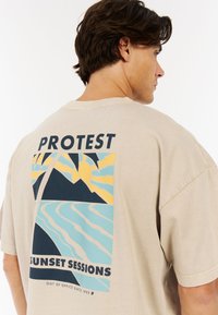 Beige oversized t-shirt met een grafische print op de achterkant van een zonsondergang, bergen, palmbomen en de tekst "PROTEST SUNSET SESSIONS."