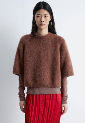 Brun fluffy kort trøje med høj hals, korte ærmer, lagdelt over en tætsiddende langærmet top, kombineret med et plisseret rødt nederdel.