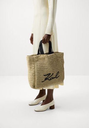 Personne portant une robe crème et des chaussures blanches à talons blocs, tenant un grand sac fourre-tout tissé beige avec des poignées noires et le nom "Karl" brodé.