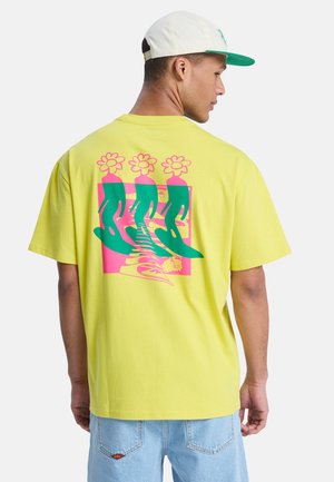Mann trägt ein gelbes T-Shirt mit einer Grafik von drei grünen abstrakten Figuren mit Blumen als Köpfen auf einem pinken Quadrat, kombiniert mit hellblauen Jeans und einer weißen Kappe.