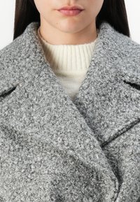Grå tekstureret frakke lavet af et blødt, fuzzy materiale og med bred krave, lagdelt over en kremfarvet ribbet sweater. Inkluderer en subtil syningdetail.