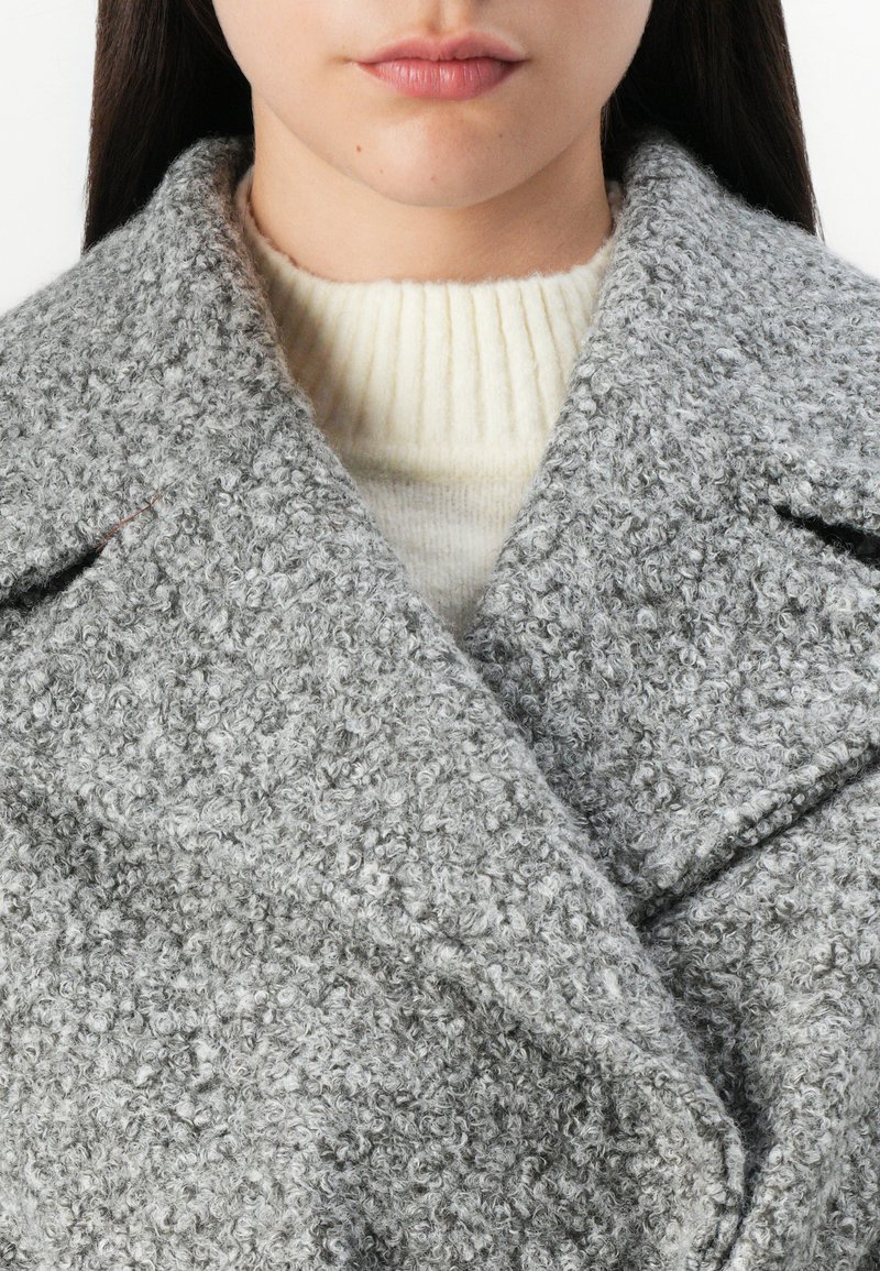 Manteau texturé gris en matériau doux et duveteux, avec un large col, superposé sur un pull côtelé crème. Comprend un détail de couture subtil.
