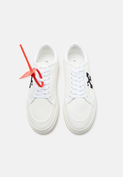 Ζευγάρι λευκά αθλητικά παπούτσια με μαύρα λογότυπα σταυρού στις πλευρές, λευκά κορδόνια και κόκκινο λουράκι ασφαλείας δεμένο στα κορδόνια του αριστερού παπουτσιού.