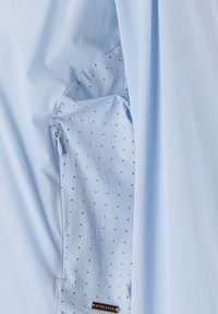 Hellblaues Jackett mit glatter Außenseite, gepunktetem Innenfutter und kleiner Reißverschlusstasche. Das Material wirkt leicht und atmungsaktiv.