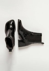 Botas negras hasta el tobillo con un acabado brillante de charol y textura de cocodrilo. Presentan un tacón ancho y paneles laterales elásticos para facilitar su uso.