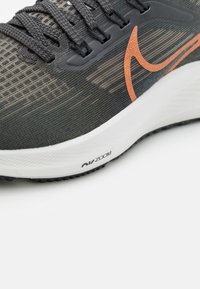 Zapatilla deportiva con una parte superior de malla de color gris oscuro con una sutil textura a rayas, logo de swoosh en naranja y una entresuela blanca marcada como "Air Zoom".