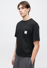 Μαύρο βαμβακερό T-shirt με κοντά μανίκια, που διαθέτει τσέπη στο στήθος και ένα πράσινο γραφικό βουνό στην τσέπη. Η σιλουέτα είναι άνετη.