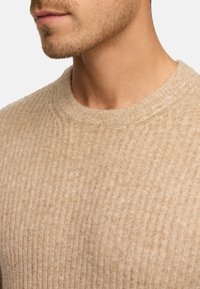 Maglione beige a coste con collo rotondo e tessuto texture, che mostra delle linee sottili. Primo piano del collo e dell'area della spalla.