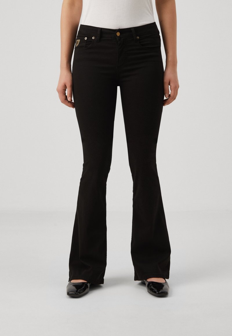 Femme portant un jean évasé noir, un haut blanc et des ballerines noires, debout devant un fond uni gris clair.