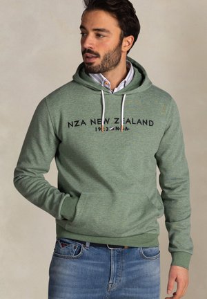 Man met een groene hoodie met de tekst "NZA New Zealand 1933", een wit shirt eronder en een blauwe spijkerbroek, die opzij kijkt.