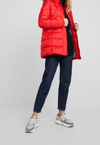 Rød pufferjakke med hætte, blank finish og kviltet design. Sammen med mørk denimjeans og multifarvede sneakers.