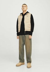 Gilet matelassé beige avec des coutures horizontales sur un hoodie noir. Un jean vert décontracté et des bottes de travail beiges complètent l'ensemble.