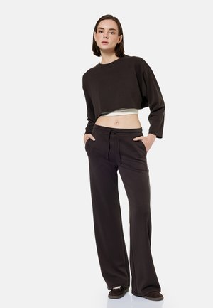 Vrouw staat met handen in de zakken, draagt een losse zwarte cropped trui, bijpassende wijde broekspijpen en donkere schoenen op een witte achtergrond.