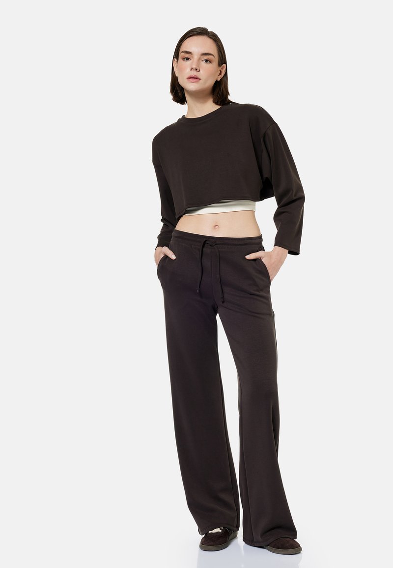 Femme debout les mains dans les poches, portant un sweat-shirt court noir ample, un pantalon large assorti et des chaussures foncées sur fond blanc.