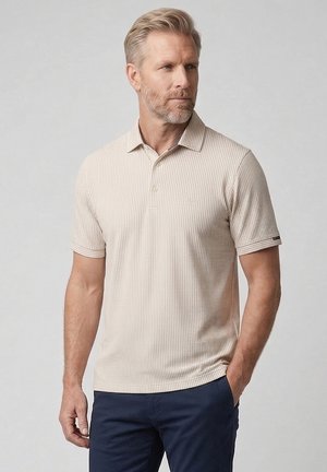 Blond man with beard wearing a beige striped short-sleeve polo shirt and navy blue pants, standing with one hand in pocket.