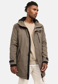 Khaki ūdensizturīga parka ar iepakojumu priekšā, regulējamu kapuci un elastīgām piedurknēm. Iespējas sānu kabatas un ar rāvējslēdzēju aizverama krūšu kabata.