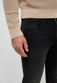 Suéter beige texturizado con puños acanalados combinado con jeans de mezclilla negros que cuentan con bolsillos frontales y un acabado liso. Mano descansando sobre los jeans.