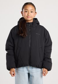 Girl wearing a black hooded puffer jacket with front pockets and light blue jeans, standing against a plain background.