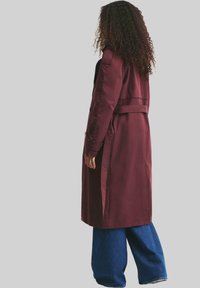 Femme aux cheveux bouclés portant un long manteau bordeaux et un jean large bleu, se tenant de côté devant un fond uni.