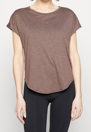 Frau trägt ein locker sitzendes braunes Kurzarmoberteil und schwarze, eng anliegende Leggings, steht vor einem schlichten hellen Hintergrund.