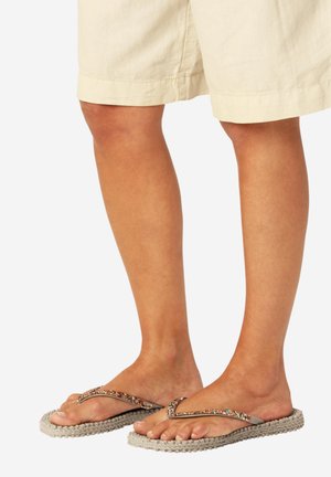 Piernas con pantalones cortos beige y chanclas beige con tiras con cuentas, de pie sobre un fondo blanco.