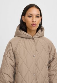 Veste à capuche matelassée beige avec un motif en losanges et une fermeture à pression, présentant une texture douce et une coupe décontractée.