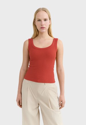 Femme aux longs cheveux blonds, portant un haut sans manches rouge côtelé et un pantalon beige taille haute, debout devant un fond uni.
