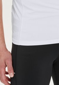 Valkoinen nopeasti kuivuva urheilu-paita verkkopaneeleilla sivuilla, yhdistetty mustiin ihonmyötäisiin legginsseihin. Lähikuva tekstuurista ja yksityiskohdista.