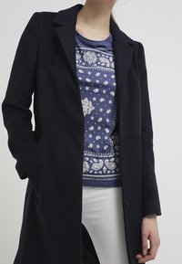 Navy ullblandad kappa med skräddarsydd design, enfärgad knapprad och sidofickor. Under finns en blå mönstrad skjorta synlig.