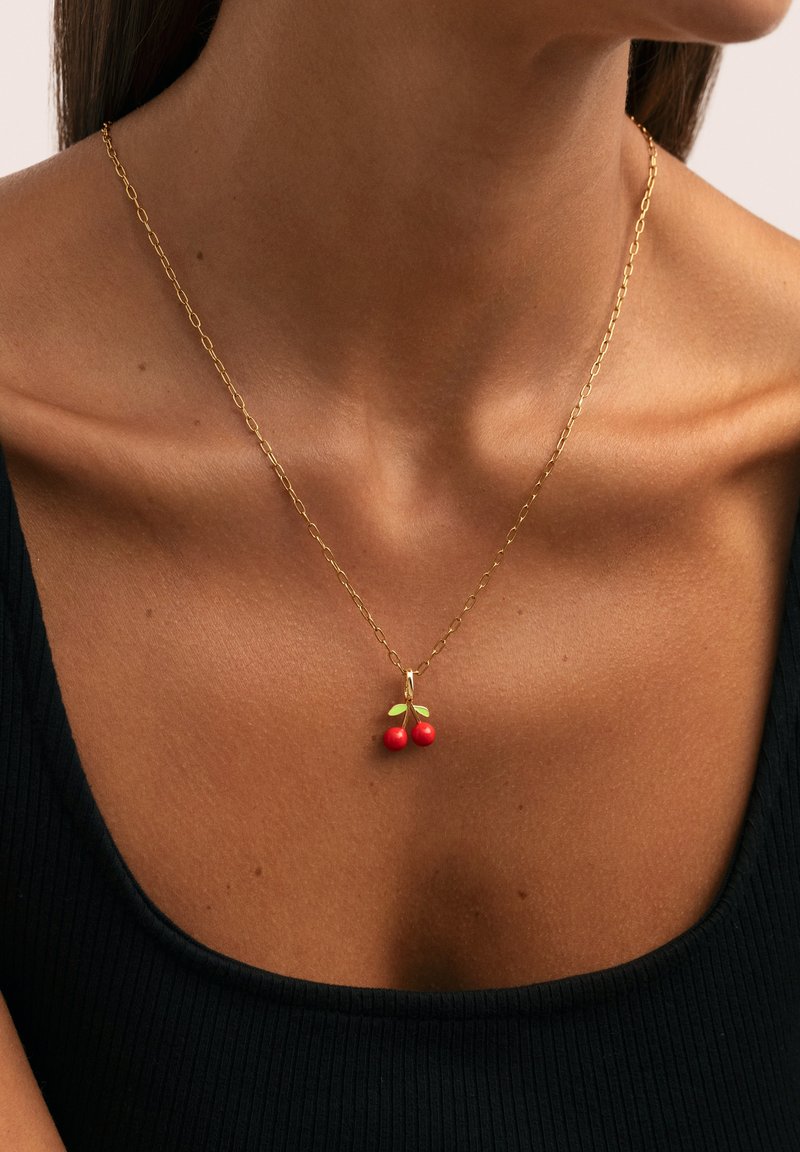 Collar de cadena de oro con un colgante de cereza que presenta dos cerezas rojas y dos hojas verdes, lucido por una mujer que lleva una blusa negra.