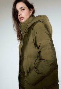 Oliwkowa kurtka puchowa z kapturem, z suwakiem z przodu, zatrzaskami i bocznymi kieszeniami. Gładka tekstura i wyściełany fason.