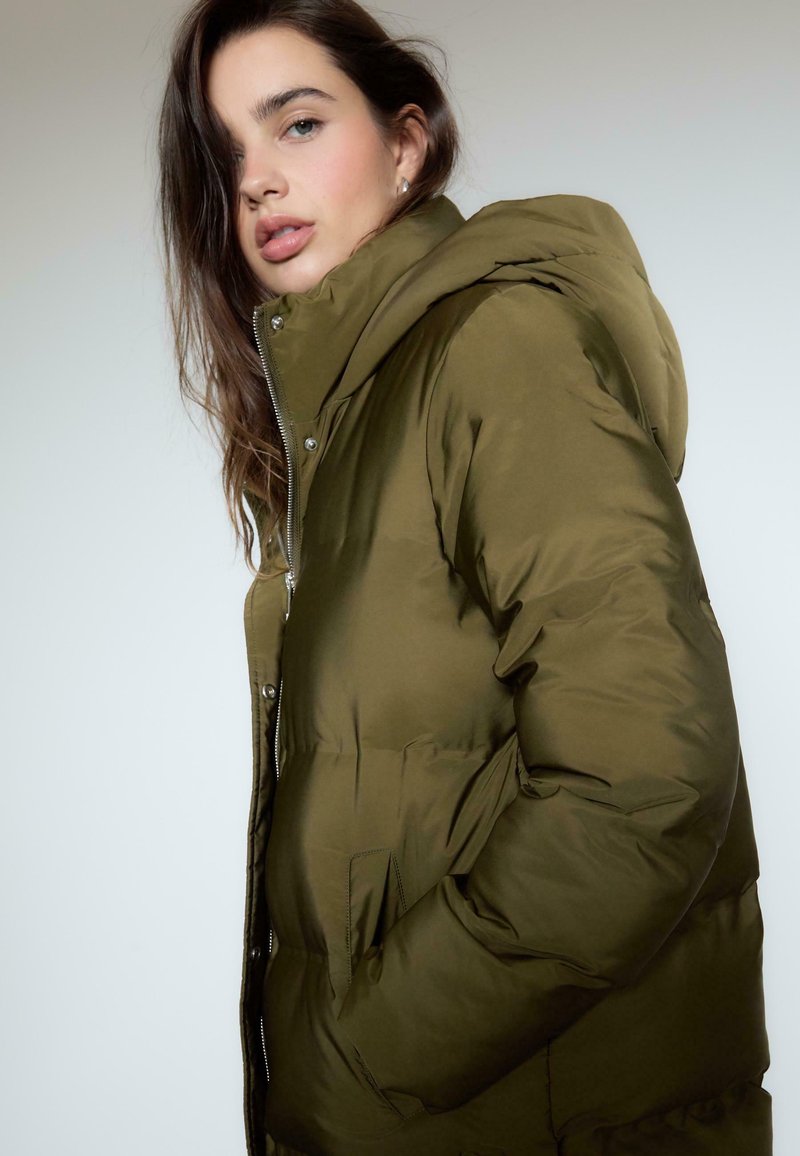 Olivově zelená péřová bunda s kapucí, která má přední zip, tiskací knoflíky a boční kapsy. Hladká textura a polstrovaný design.