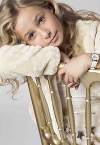 Enfant reposant sur un garde-corps peint en or, portant un doux manteau en fourrure couleur crème, avec des cheveux blonds ondulés et une montre en argent au poignet.