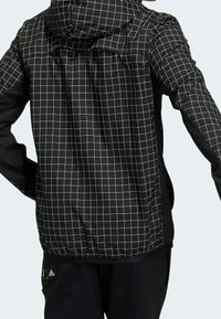 Musta takki, jossa on valkoinen ruutukuosi, hupulla ja nyörillä. Valmistettu sileästä materiaalista, suunniteltu istuvaksi.