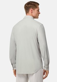 Camisa de manga larga gris claro con cuello abotonado, textura suave, dobladillo recto y puños abotonados, vista desde la parte de atrás.