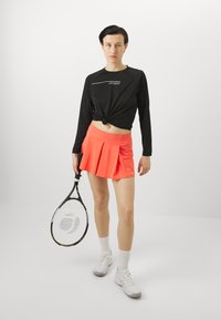 Crna majica dugih rukava zavezana u struku, u kombinaciji s nabranom koraljno ružičastom tenis suknjom. Drži reketa, nosi bijele čarape i cipele.