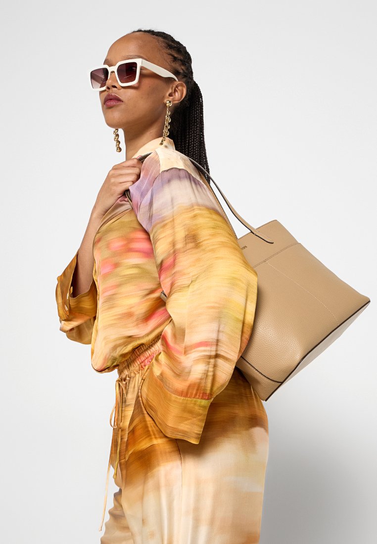 Donna con capelli intrecciati che indossa occhiali da sole bianchi, orecchini d'oro, una blusa multicolore e pantaloni, mentre tiene una grande borsa beige a tracolla.